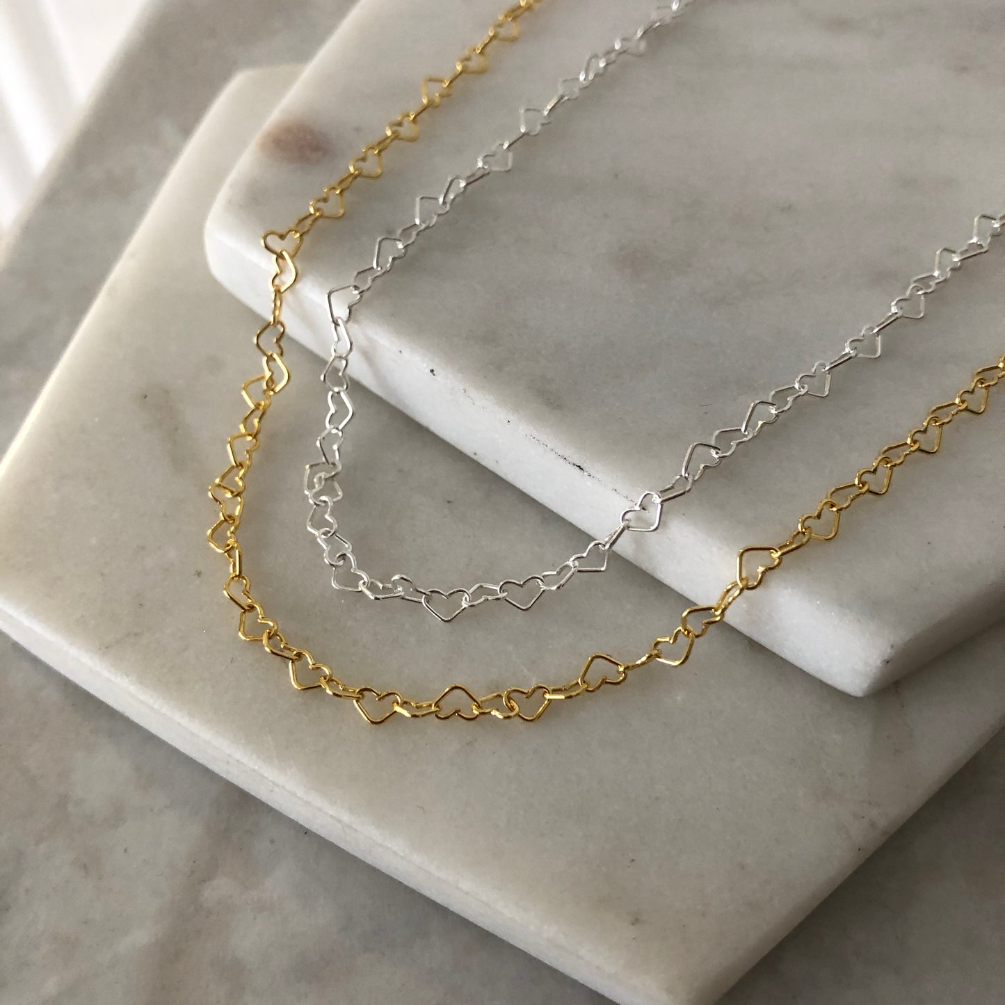 Two heart chain necklaces, one gold-colored and one silver-colored, laid on a white marble surface.