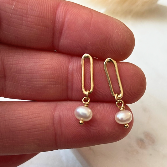 Gold earrings with pearl drops held between fingers on a neutral background