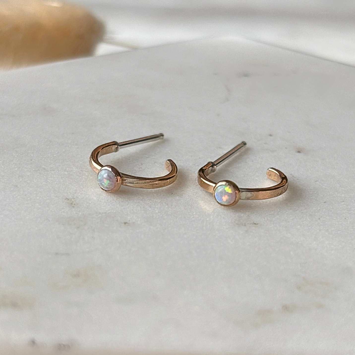 A pair of small gold huggie hoop earrings with round iridescent opal gemstones, resting on a light marble surface.