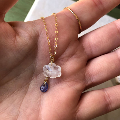 A hand holding a rainbow moonstone cloud pendant with a purple tanzanite teardrop.