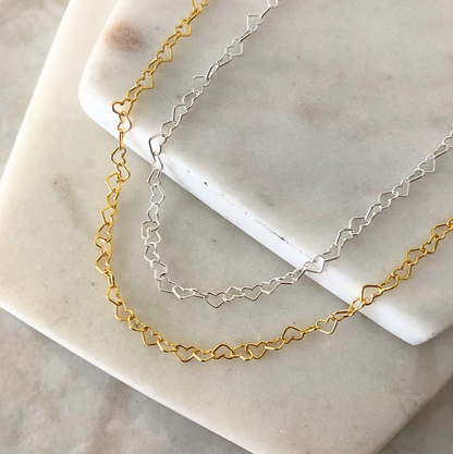 Two heart chain necklaces, one gold-colored and one silver-colored, laid on a white marble surface.