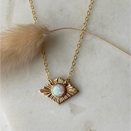 A gold-toned necklace with a textured, diamond-like pendant featuring a central iridescent opal gemstone, displayed on a light marble surface with a beige dried flower stem.