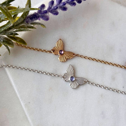 Two butterfly bracelets, one with a gold fill chain and rose-cut tanzanite gemstone, and the other with a silver chain and a similar gemstone, displayed on a white surface next to a purple flower.