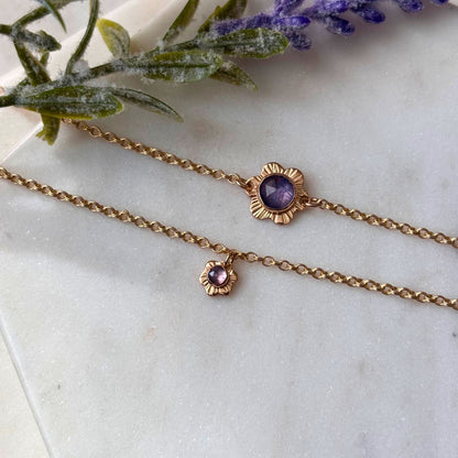 Two gold-filled bracelets with floral details and rose-cut tanzanite gemstones on a marble surface, with a small purple flower arrangement in the background.