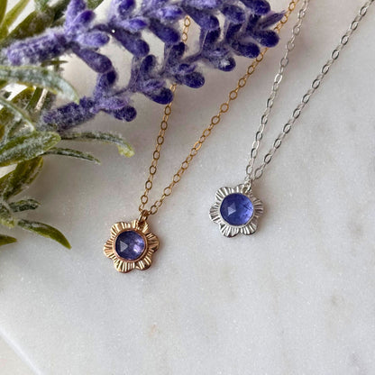 A collection of flower necklaces with varying sizes, made of 14k gold-fill or sterling silver, featuring a central tanzanite gemstone, displayed against a marble background with floral elements.