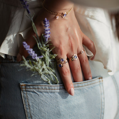 A woman's hand featuring gold flower rings and a sprig of lavender, reaching into her pocket.