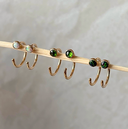 Four gold hoop earrings with round gemstones are displayed on a wooden stick against a soft, light gray fabric background. Two earrings feature iridescent white opal-like stones, and two feature deep green stones.