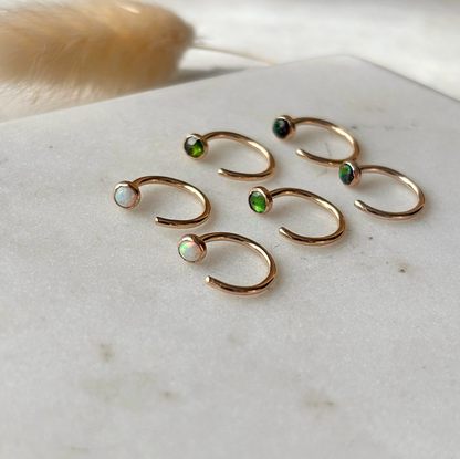 Six gold open hoop earrings with small iridescent gemstone accents in green, blue, and white, arranged on a light marble surface.