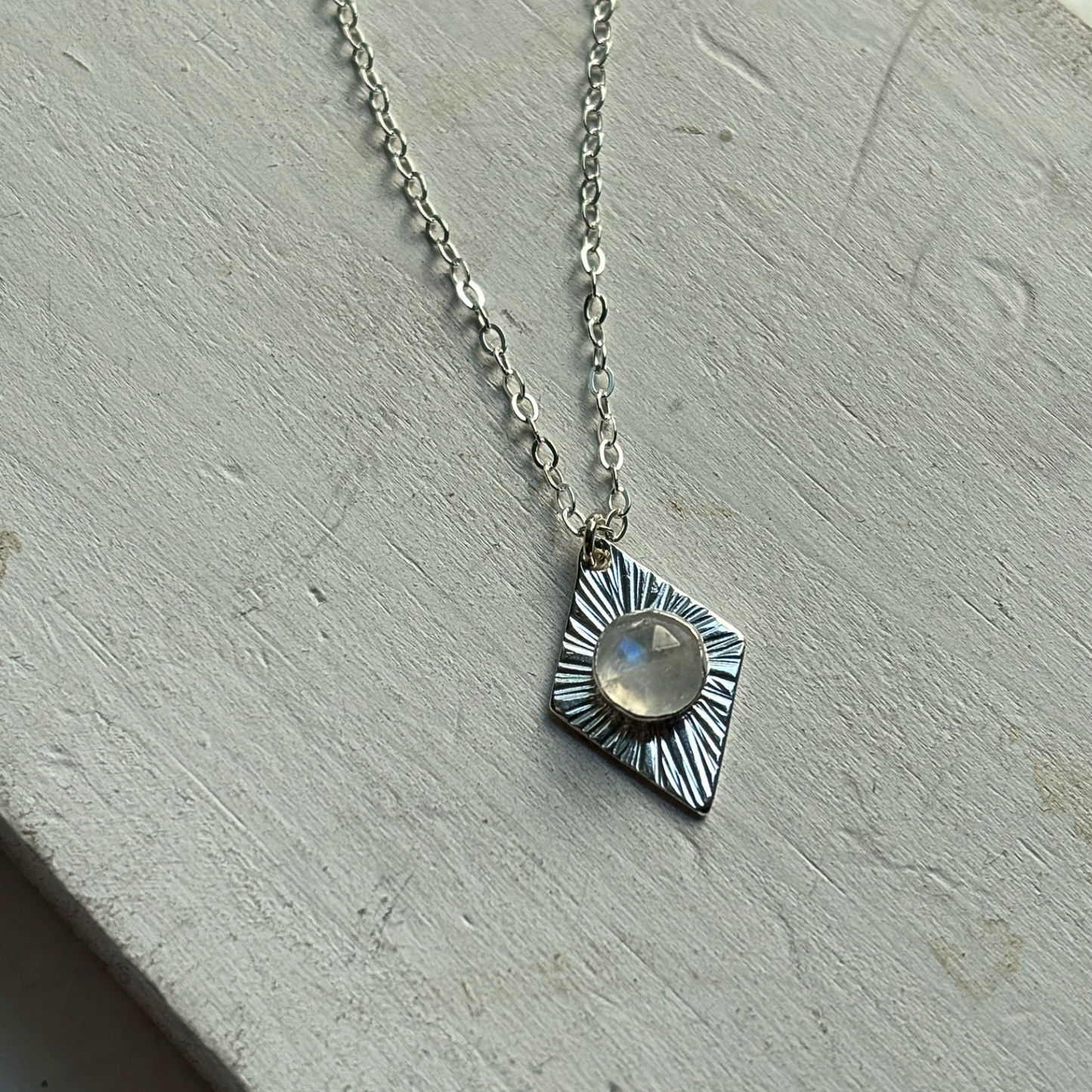 A silver necklace with a textured, diamond-shaped pendant featuring a central round clear stone, displayed on a light gray textured surface.