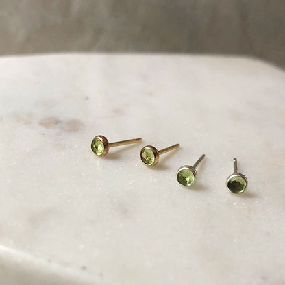 A set of small round stud earrings made with peridot stones, displayed on a marble surface.