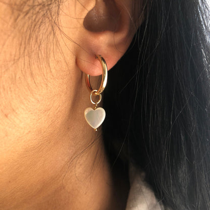 A Mother of Pearl charm heart dangling from a gold hoop earring.