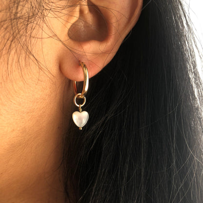 A Mother of Pearl charm small heart dangling from a gold hoop earring.