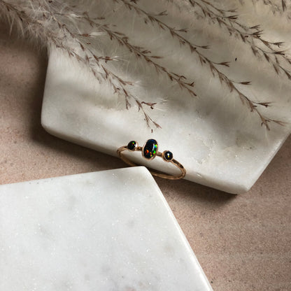 An oval triple gemstone ring with black opal set in gold displayed on a white surface.