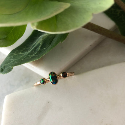 An oval triple gemstone ring with black opal set in gold displayed on a white surface.