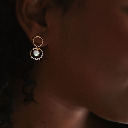 A person with brown skin wearing a pair of mixed metal prism gold circle opal drop earrings with sterling silver textures.