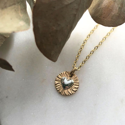 A gold-filled necklace with a textured circular pendant featuring a puffed heart in the center, displayed on a white surface with a leaf in the background.