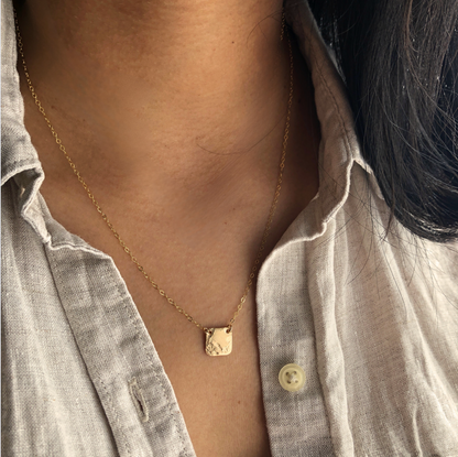 A square pendant necklace with a textured surface worn by a person with a partial view of their neck.