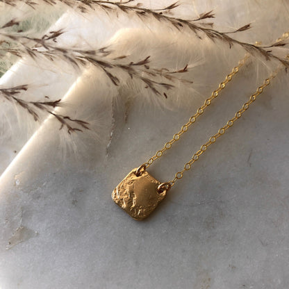 A square textured gold pendant on a marble surface.