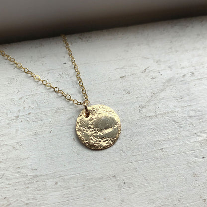 A gold medallion necklace with a textured surface laying on a light surface, with the chain visible.