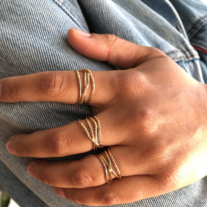 A hand wearing multiple gold rings with multiple criss crossing wires on a neutral background.