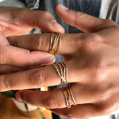 A hand wearing gold rings with multiple criss crossing wires on a neutral background.