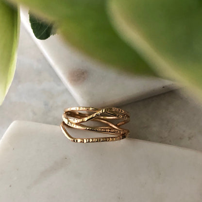 A gold-filled ring with a crisscrossing texture, placed on a marble surface partially visible behind some green leaves.