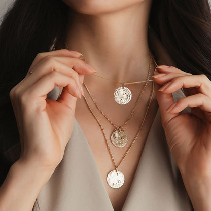A gold zodiac pendant on a chain with other zodiac pendants in the background, being held up by fingers.