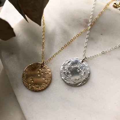 Two zodiac pendant necklaces, one with a gold-fill pendant and the other with a silver pendant, both on a cable chain, displayed against a neutral background.