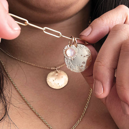 A gold zodiac pendant on a chain with a moonstone pendant, being held up by fingers.